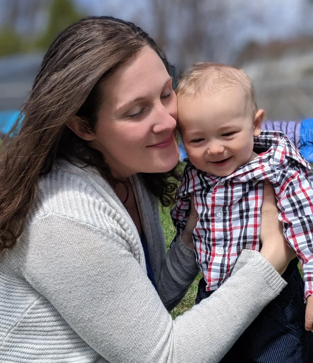 A woman with long brown hair and a light sweater gently holding a smiling young boy with short blonde hair and a plaid shirt outdoors.