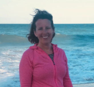Woman smiling at the beach with ocean waves in the background, wearing a pink zip-up jacket and a striped top.