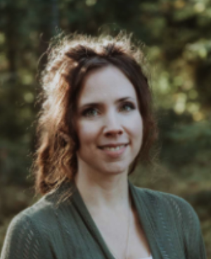 A woman with brown curly hair smiling outdoors with trees in the background.