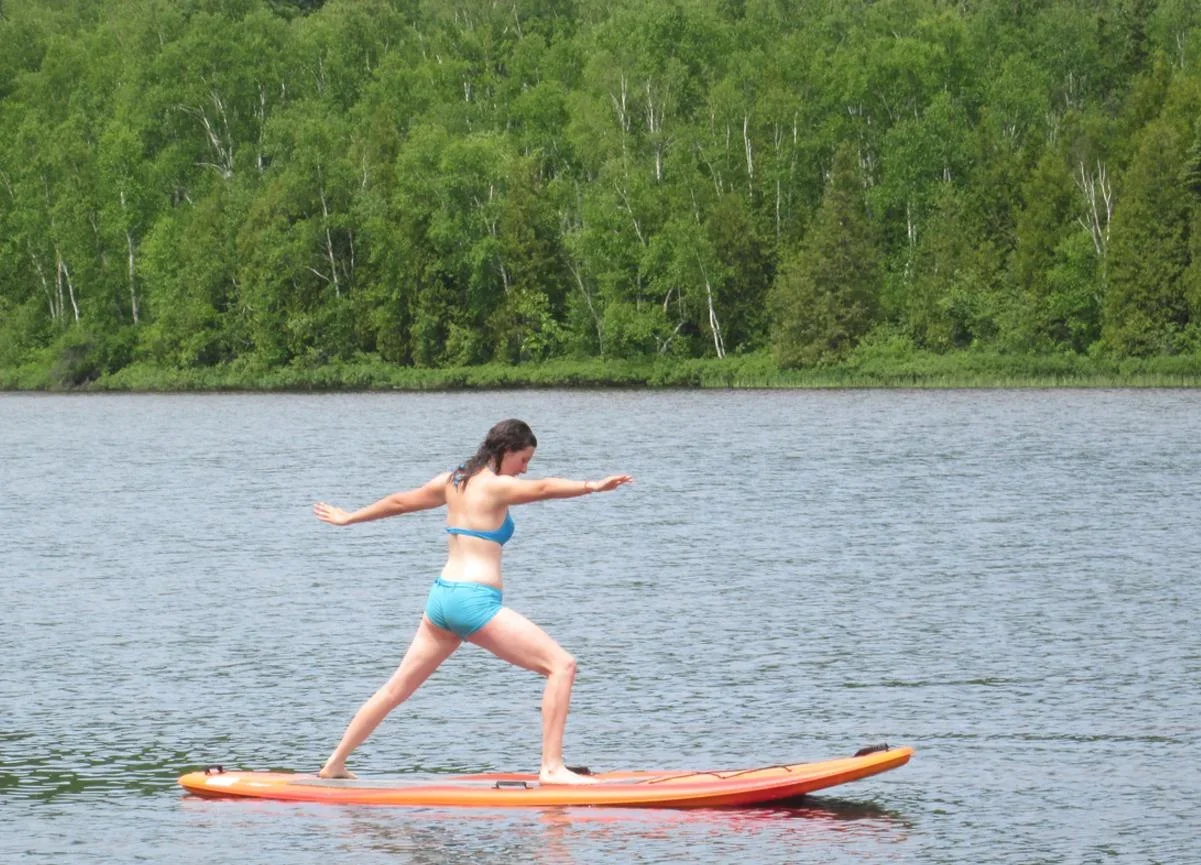 Sandie Gascon in a blue bikini balancing on an orange paddleboard on a lake with green trees in the background.