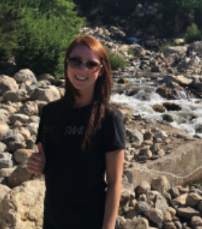 A woman with red hair wearing sunglasses and a black T-shirt standing by a rocky stream with greenery in the background.