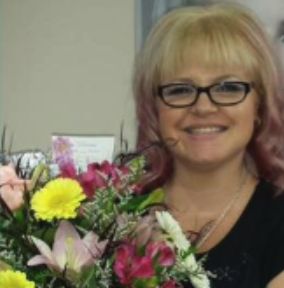 A woman with blonde hair and glasses smiling while holding a large bouquet of colorful flowers.