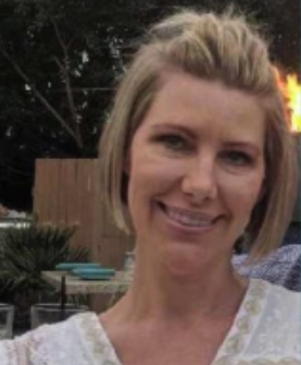 A woman smiling outdoors with a wooden fence, trees, and a firepit in the background.