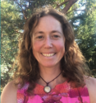 A woman with curly brown hair smiling outdoors, wearing a pink and purple dress and a necklace with a round pendant, surrounded by trees and sunlight.