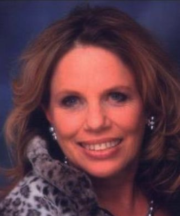 A woman with light brown, wavy hair smiling, wearing pearl earrings and a leopard-print fur coat.