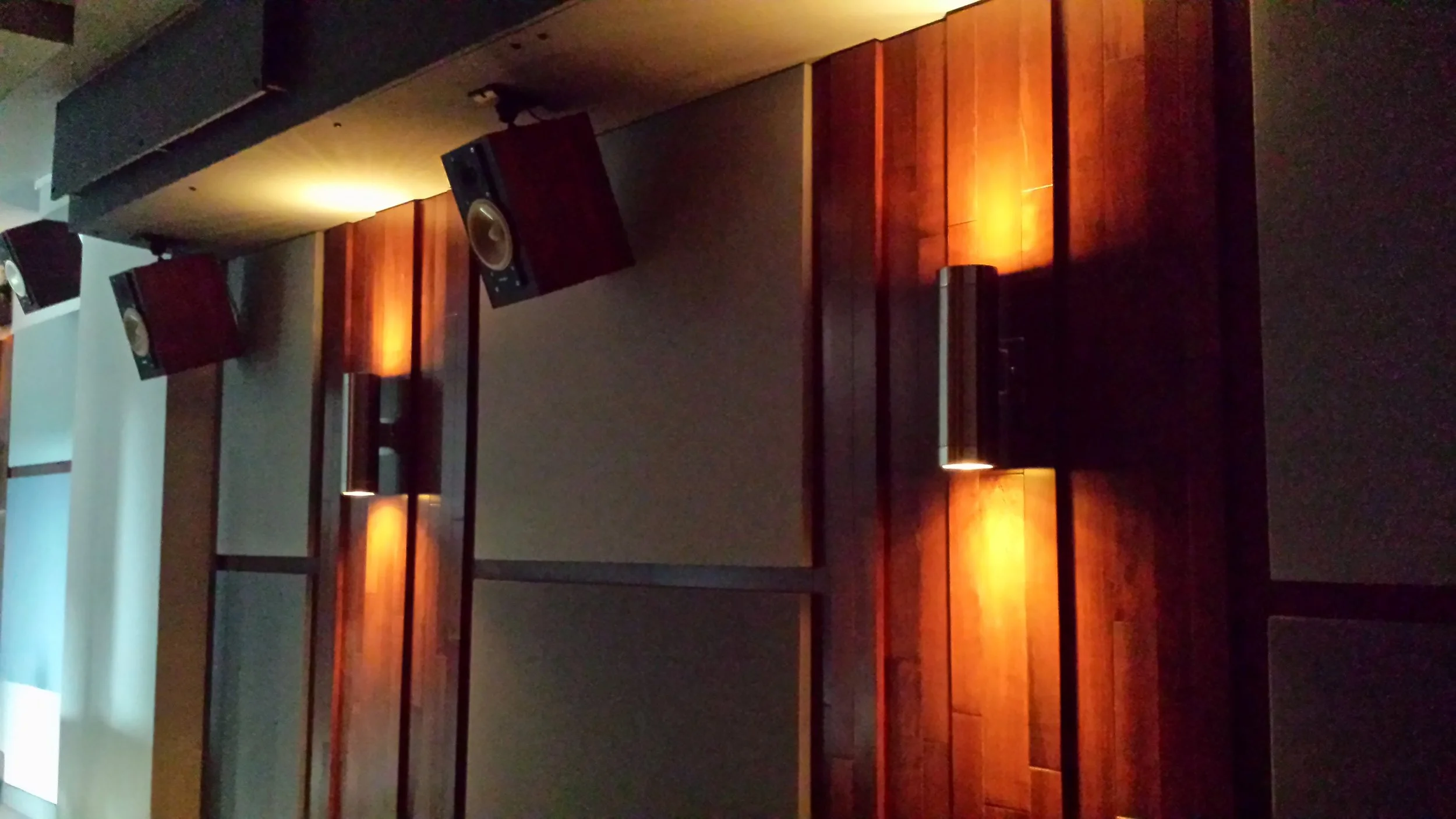 A dark auditorium with wooden walls and acoustic panels, featuring two suspended speakers and dim lighting.