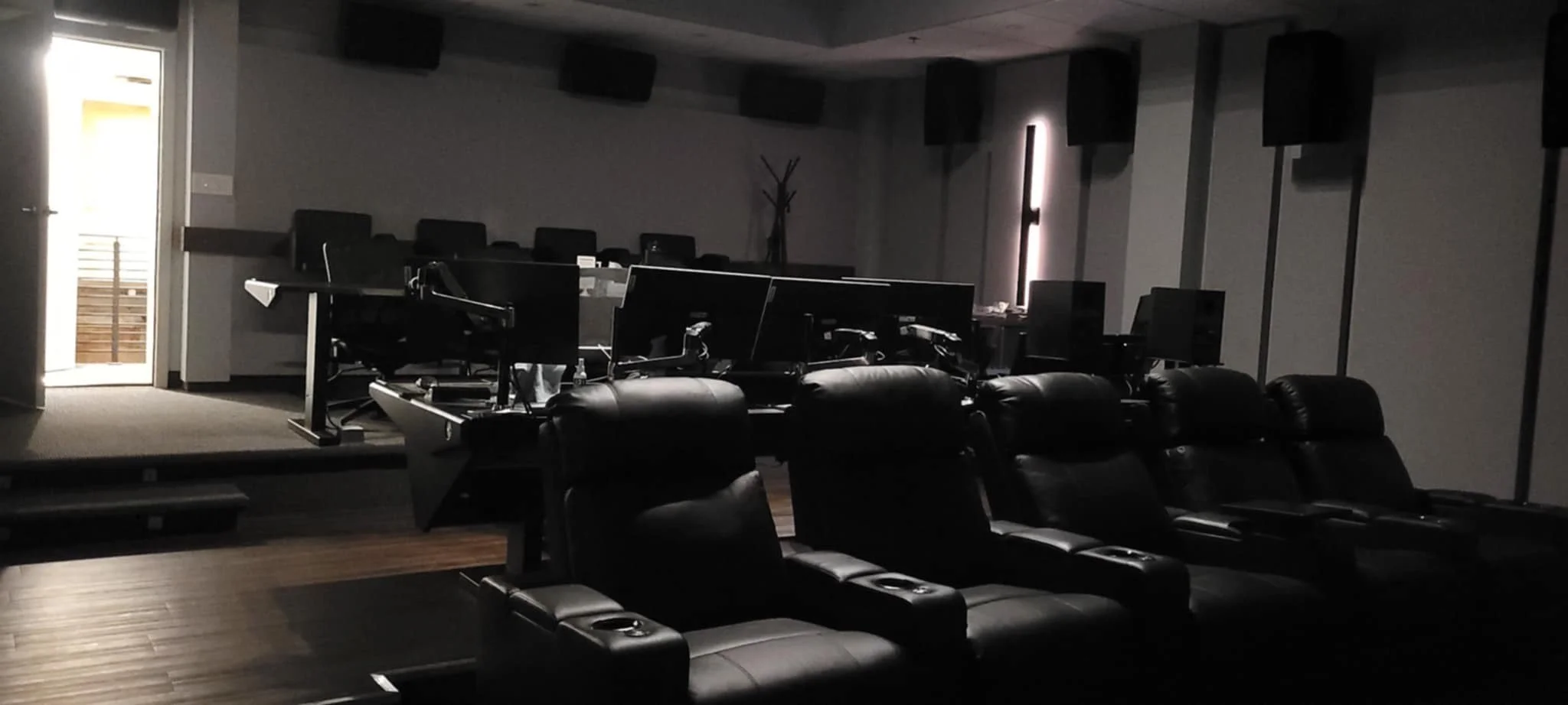 Pièce sombrement éclairée avec des sièges de cinéma en cuir noir et plusieurs écrans d’ordinateur sur les bureaux, probablement une salle de jeu ou de visionnement de films, avec une porte donnant sur un balcon ou un espace extérieur à gauche.