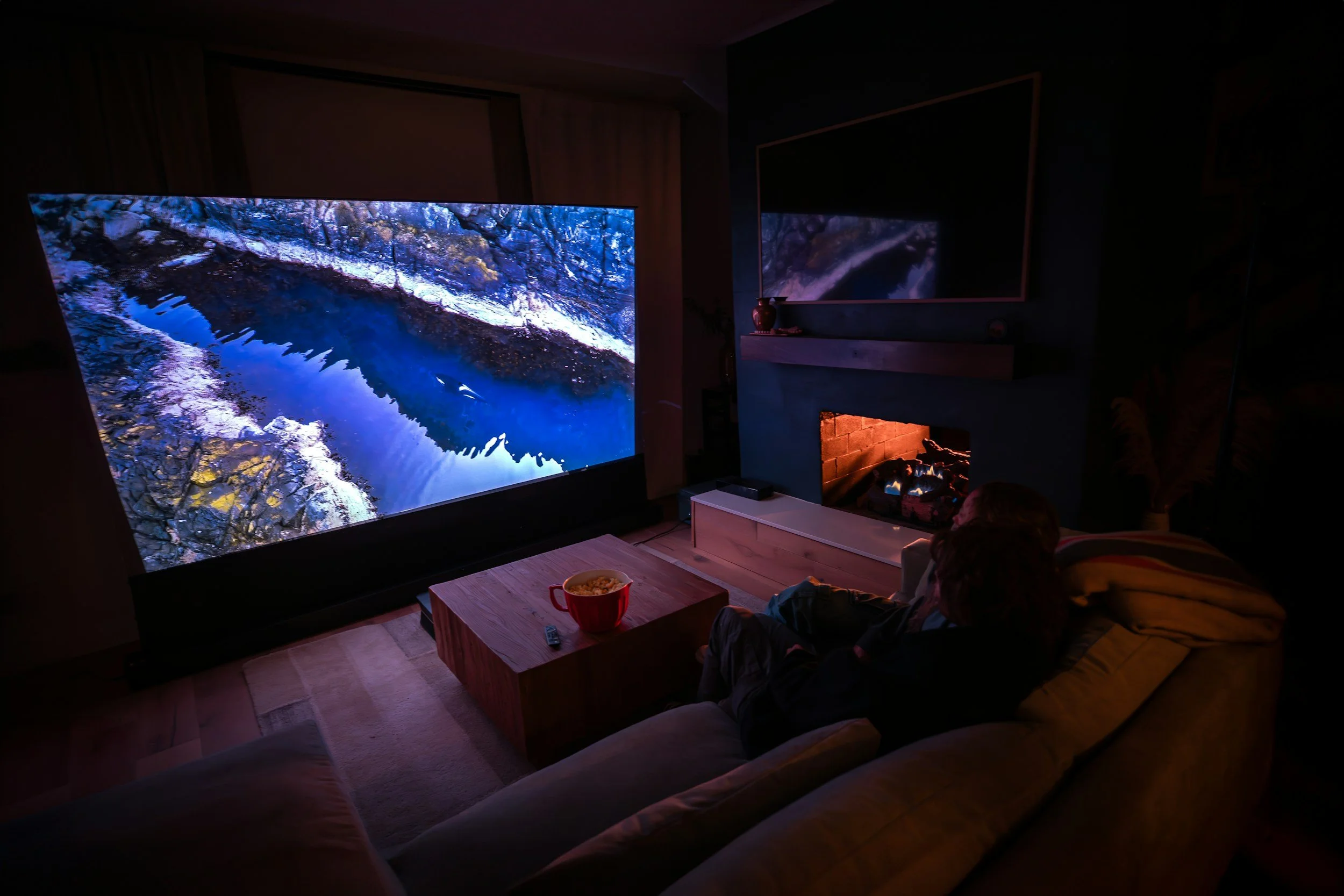 A person sitting on a beige couch watching a large television screen displaying a nature scene of a river or stream surrounded by snowy and rocky terrain. The room has a fireplace with a small fire burning, a wooden coffee table with a bowl of popcorn and TV remote, and a dark wall with a mounted television or mirror reflecting the scene.