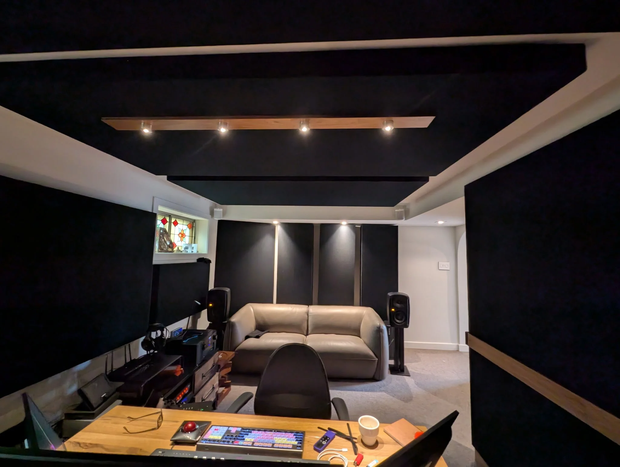 A cozy music or recording studio with black acoustic panels on the walls and ceiling. A small window with stained glass is on the left, and a beige sofa is against the back wall. There are two speakers on stands, a mixing desk, and electronic equipme