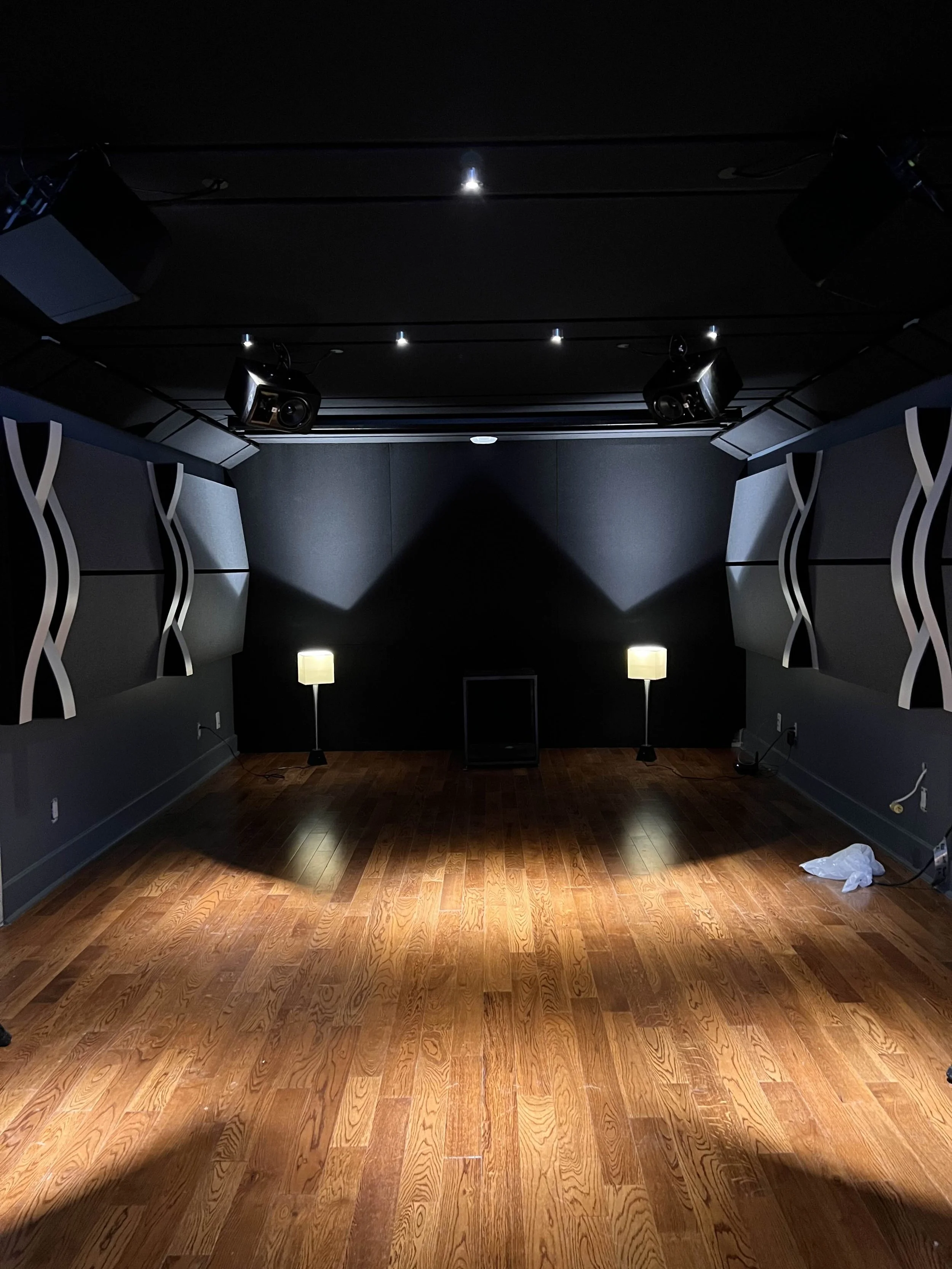 Empty dark room with wood floor, two lamps, and black acoustic wall panels, with stage lighting equipment.