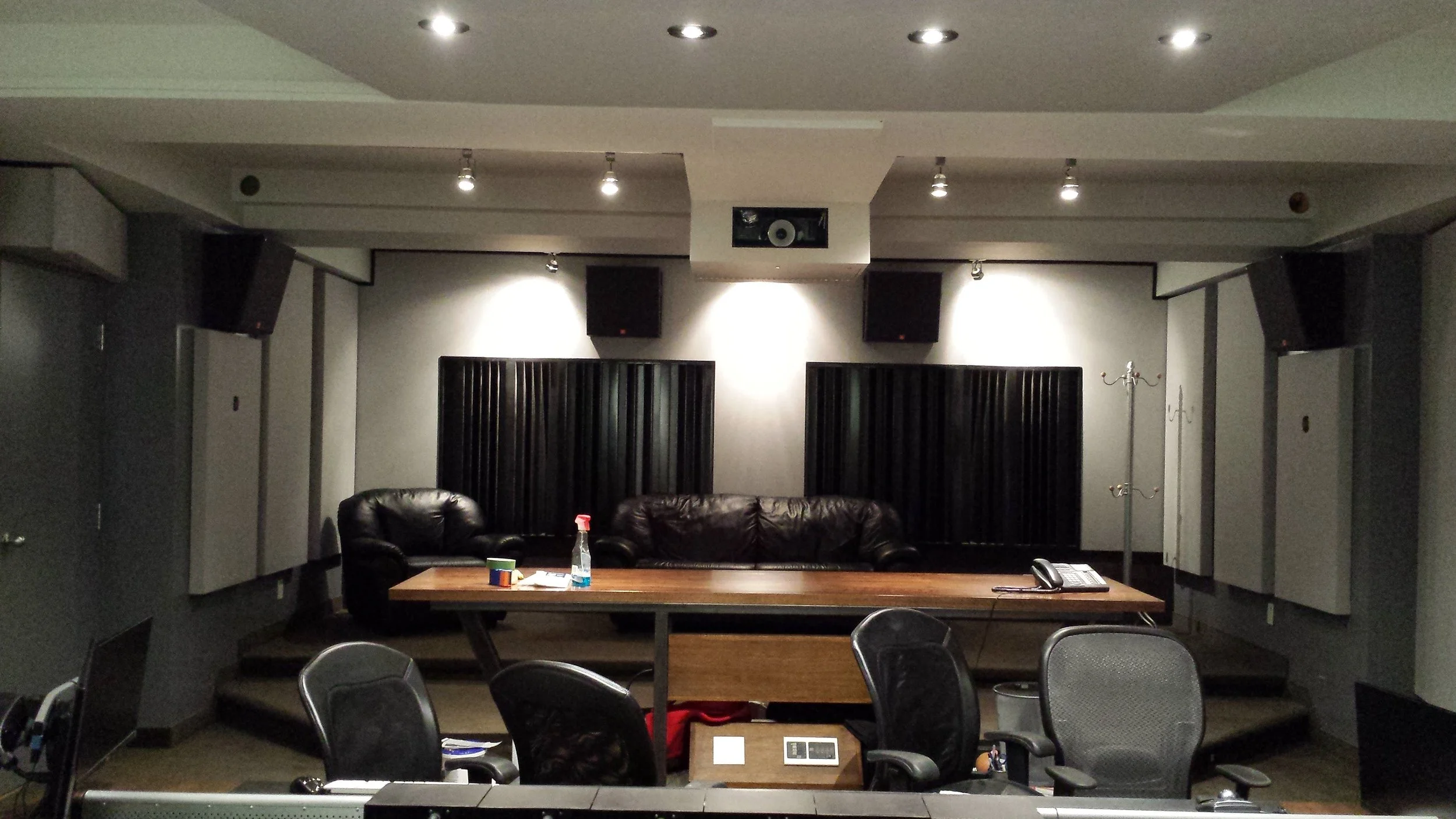 A recording studio or conference room with a stage, black leather sofas, and a table with a spray bottle, a cup, and some papers. There are black acoustic panels on the back wall, multiple ceiling lights, and several chairs in the foreground. The roo