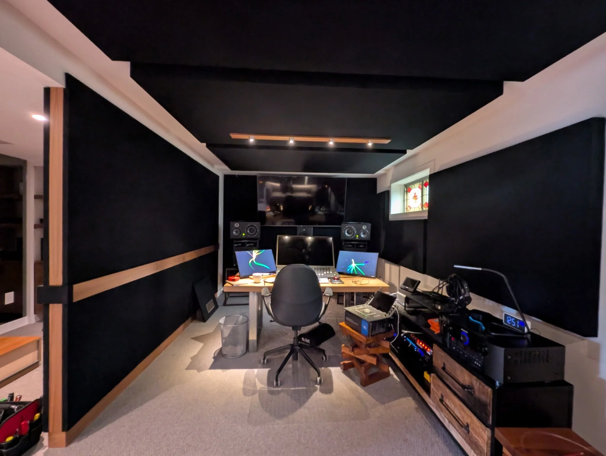 A professional music or recording studio with black acoustic panels, two computer monitors, studio speakers, audio mixing equipment, and a black office chair in the center.