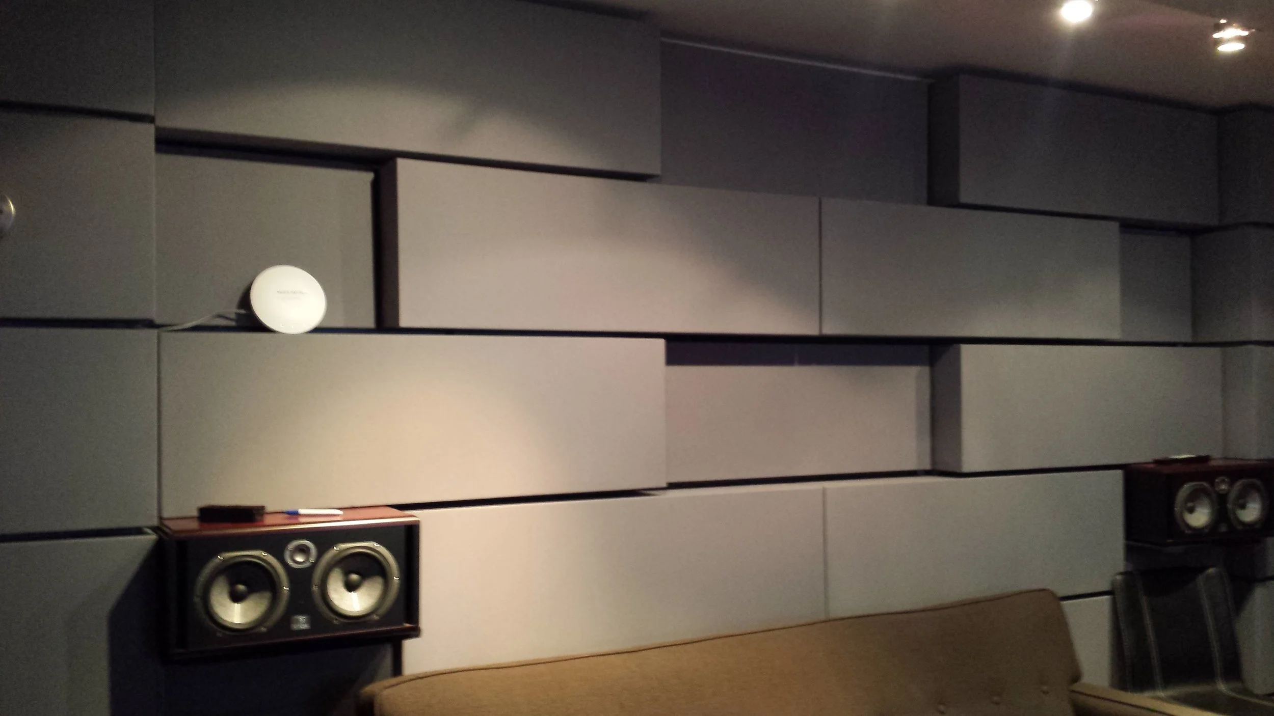 A wall with gray acoustic panels, two black speaker cabinets, and a small white round electronic device connected with wires. There is a brown couch and black chair in the foreground.