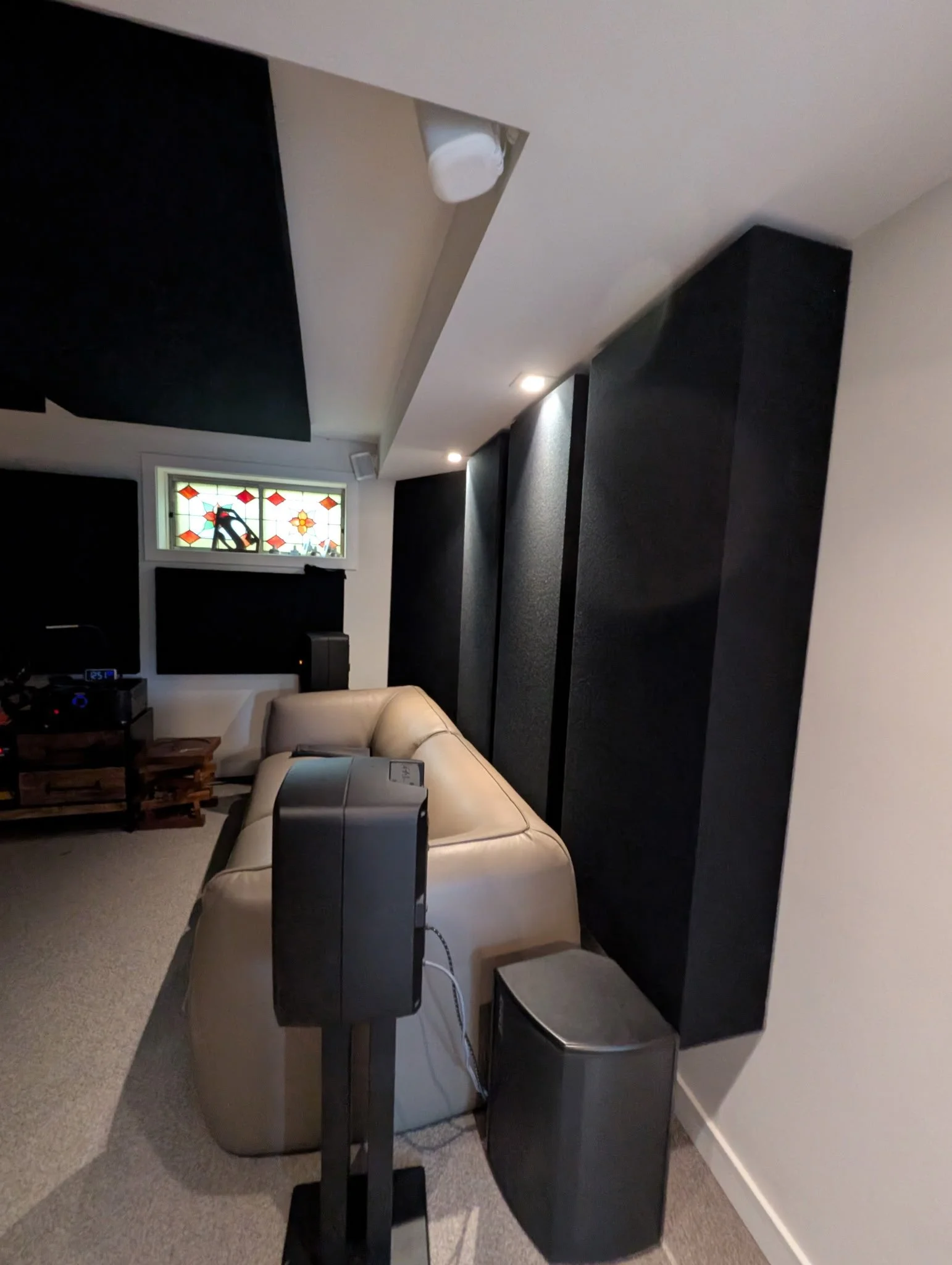 A home theater room with black acoustic panels on the walls, a beige leather sofa, a small window with stained glass, and audio equipment including a speaker, a subwoofer, and a stereo receiver.