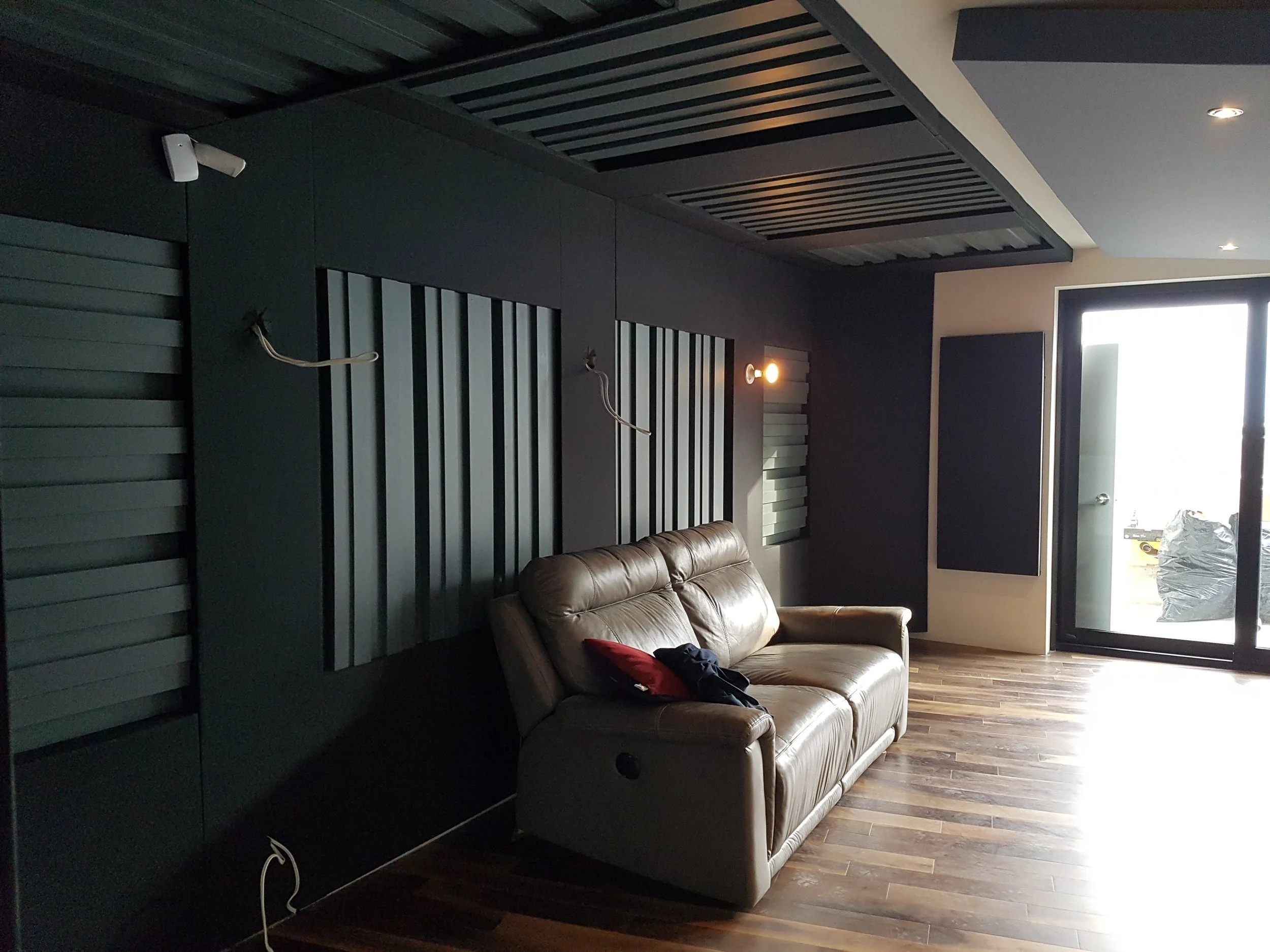 Living room with dark wall panels, a beige leather sofa, wooden flooring, and a glass door leading outside. The wall has two light fixtures and some exposed wires.