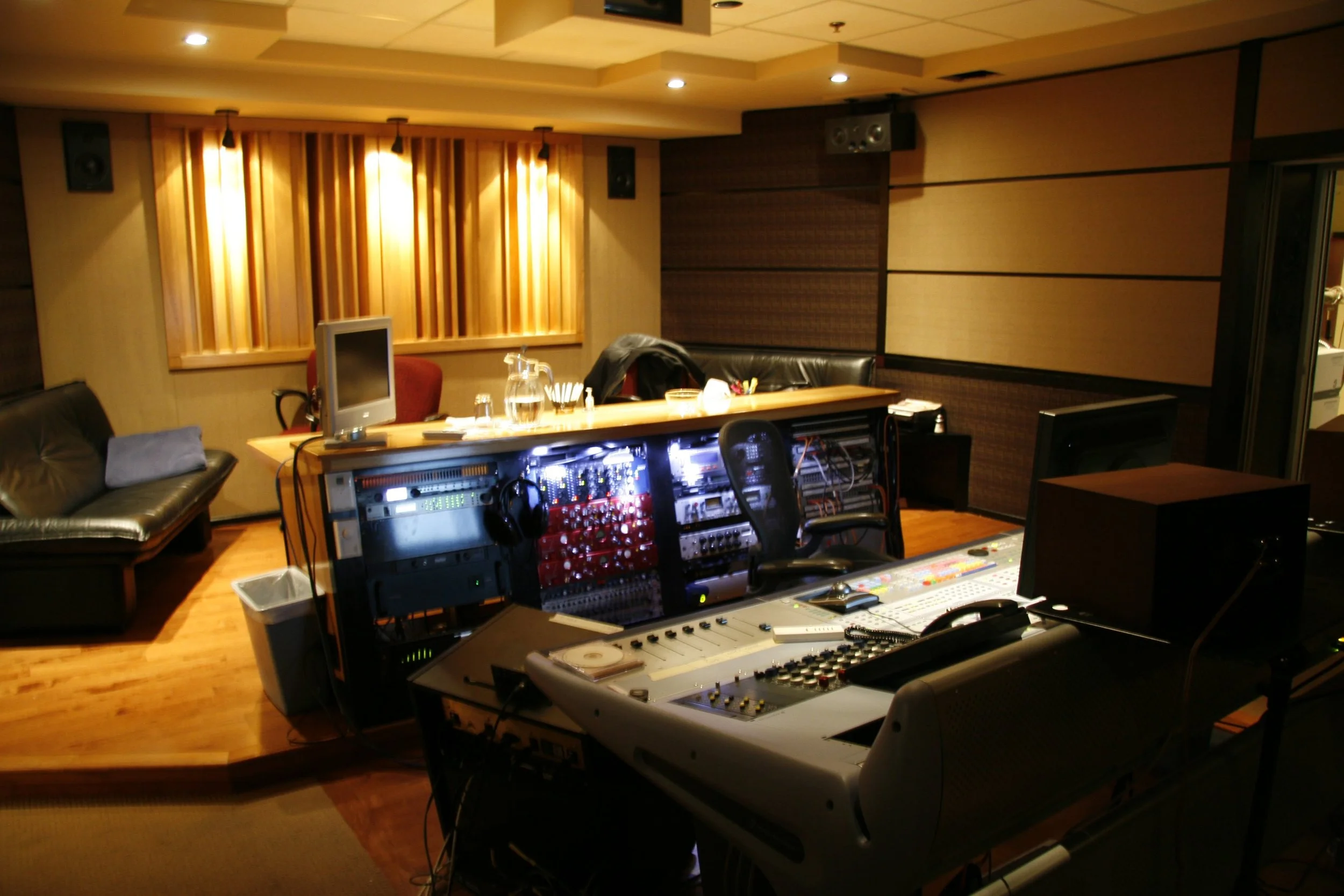 Recording studio with mixing console, audio equipment, computer monitors, and a black sofa in a wood-paneled room.