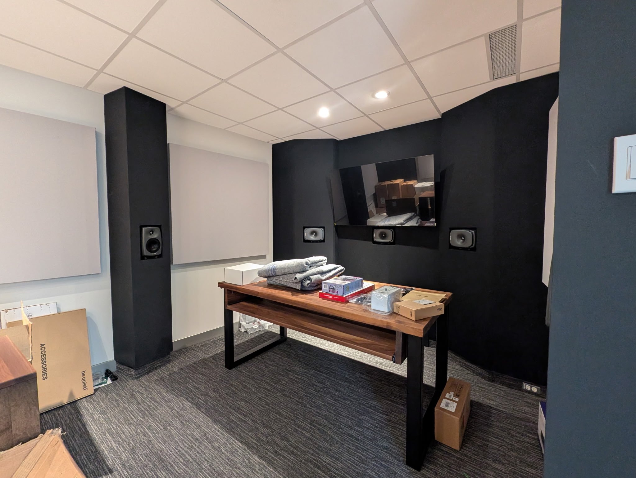 An interior room with a wooden table holding clothes and boxes, black walls with mounted speakers, painted soundproofing panels, and a large flat-screen TV on the wall.