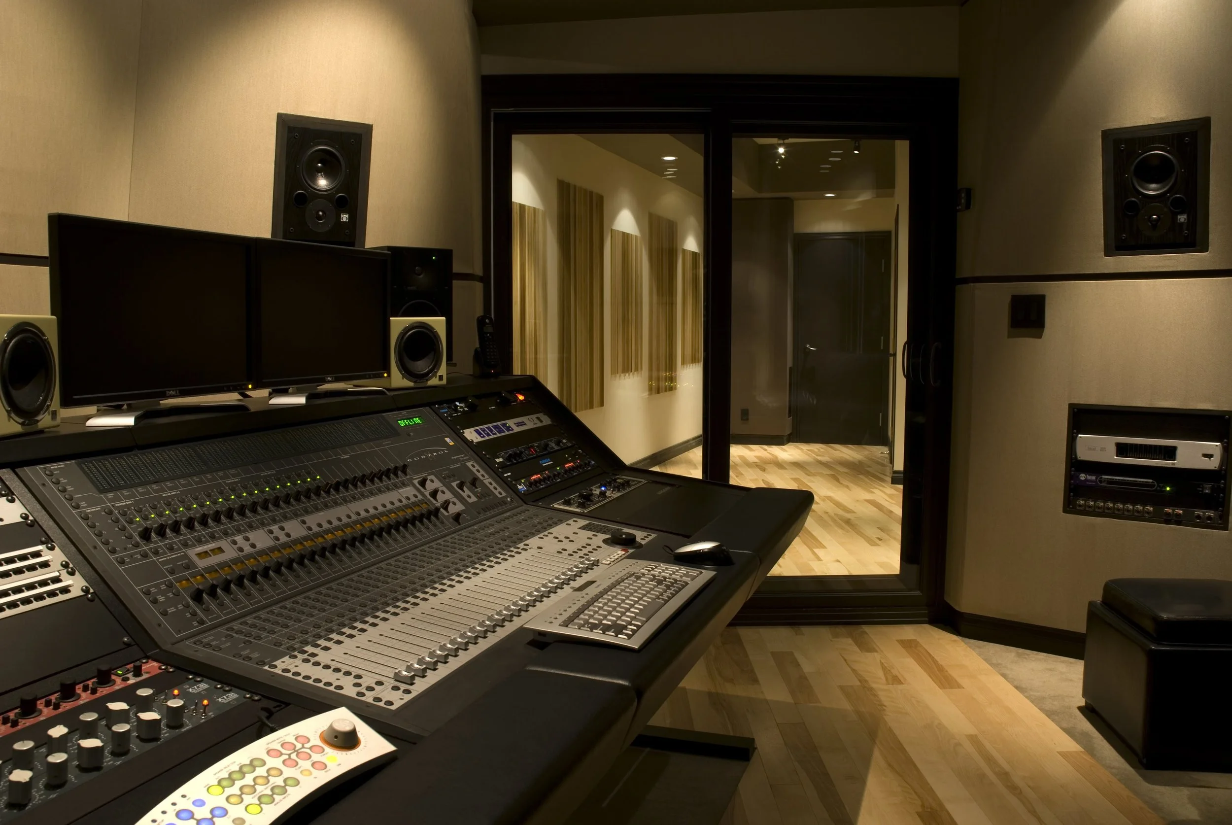 A professional recording studio control room with a large mixing console, multiple monitors, and various audio equipment. There are acoustic panels on the walls and a door leading to another room.