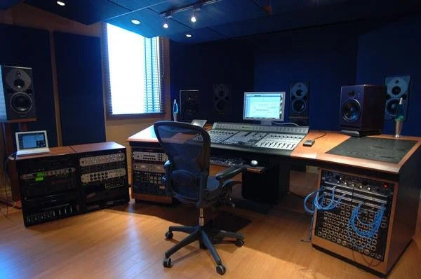 Audio recording studio with mixing console, studio monitors, rack-mounted equipment, a computer monitor, and a swivel chair in a professional setting.