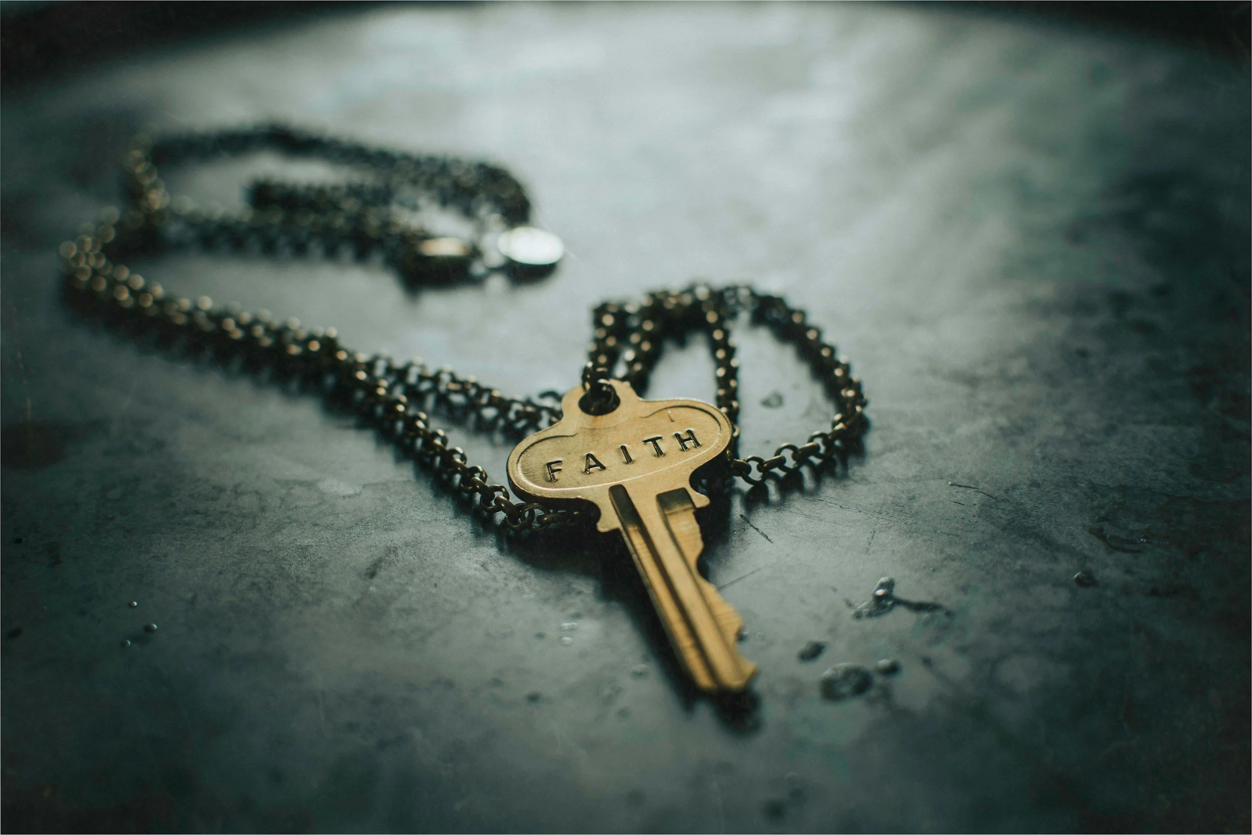 A close-up of a pendant necklace with a key and heart-shaped tag engraved with the word 'FAITH' on a dark, textured surface.