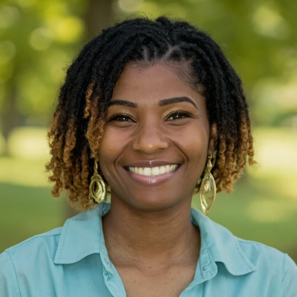 A smiling woman with short dreadlocks and gold earrings, wearing a blue collared shirt, outdoors with green trees in the background.