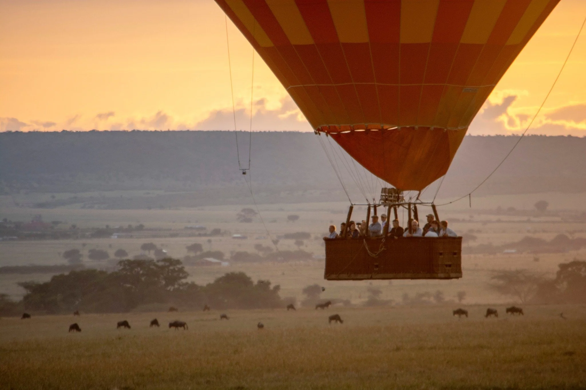 A hot air balloon carrying travelers floats above the Serengeti plains during the great wildebeest migration, offering a breathtaking safari view by Orange Moon Travel.