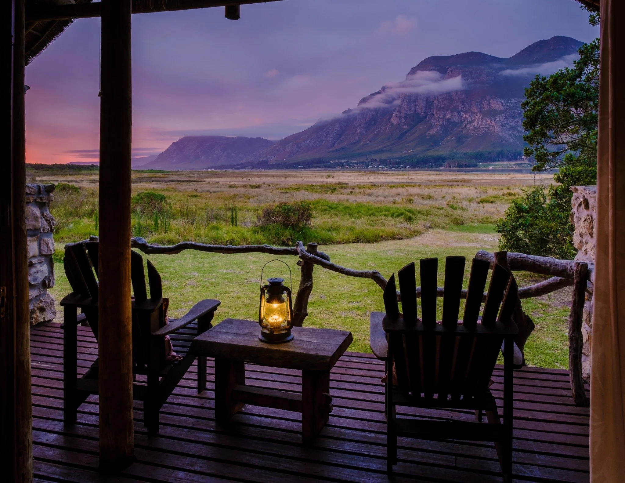 A luxury wooden lodge with chairs overlooking mountains and grasslands near Hermanus on the Garden Route, Western Cape, South Africa, during sunset — by Orange Moon