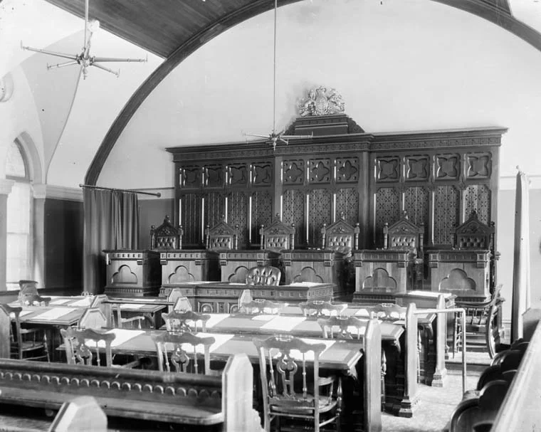 Empty courtroom in black and white featuring traditional wooden seating, a grand judge’s bench, and vaulted ceiling with hanging lights — symbolizing justice, legal expertise, and the professional environment of Melid Hasaj Legal Firm.