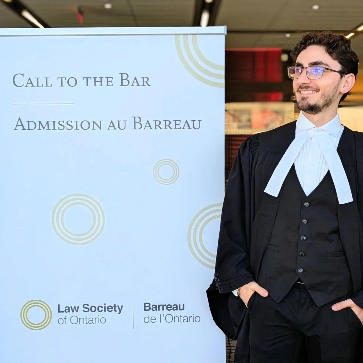 A man dressed in legal robes standing next to a sign that reads 'Call to the Bar / Admission au Barreau' with logos of the Law Society of Ontario. He is smiling, wearing glasses, and has his hands in his pockets.