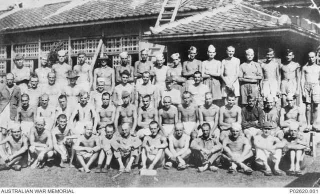 Black and white historical photograph of a group of shirtless men, some wearing hats and shorts, standing and sitting outdoors in front of a large building with a tiled roof and a ladder leaning against it, at the Australian War Memorial.