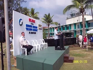 Outdoor event with a stage, palm trees, and New Zealand flags, featuring a speaker at a podium, a seated man, and several empty chairs.