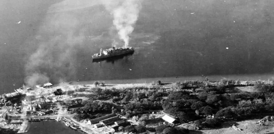 Black and white aerial photo of a coastal town with a ship emitting smoke near the shoreline.