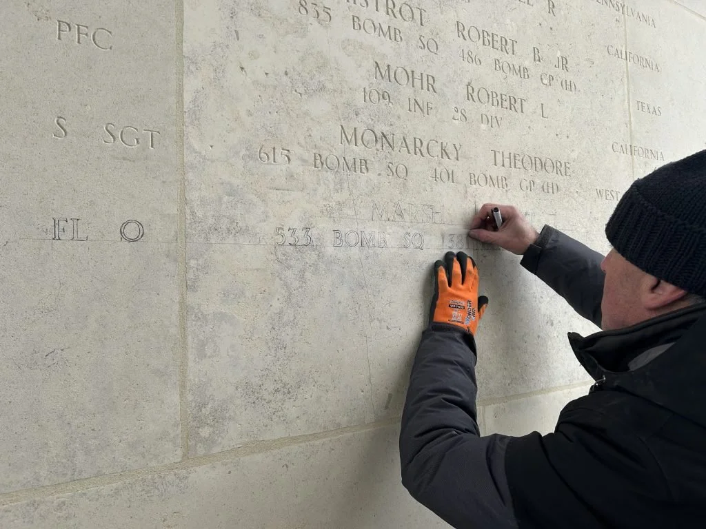 A person wearing a black beanie, black jacket, and orange gloves is carving or cleaning engraved names and information on a memorial wall made of light-colored stone.