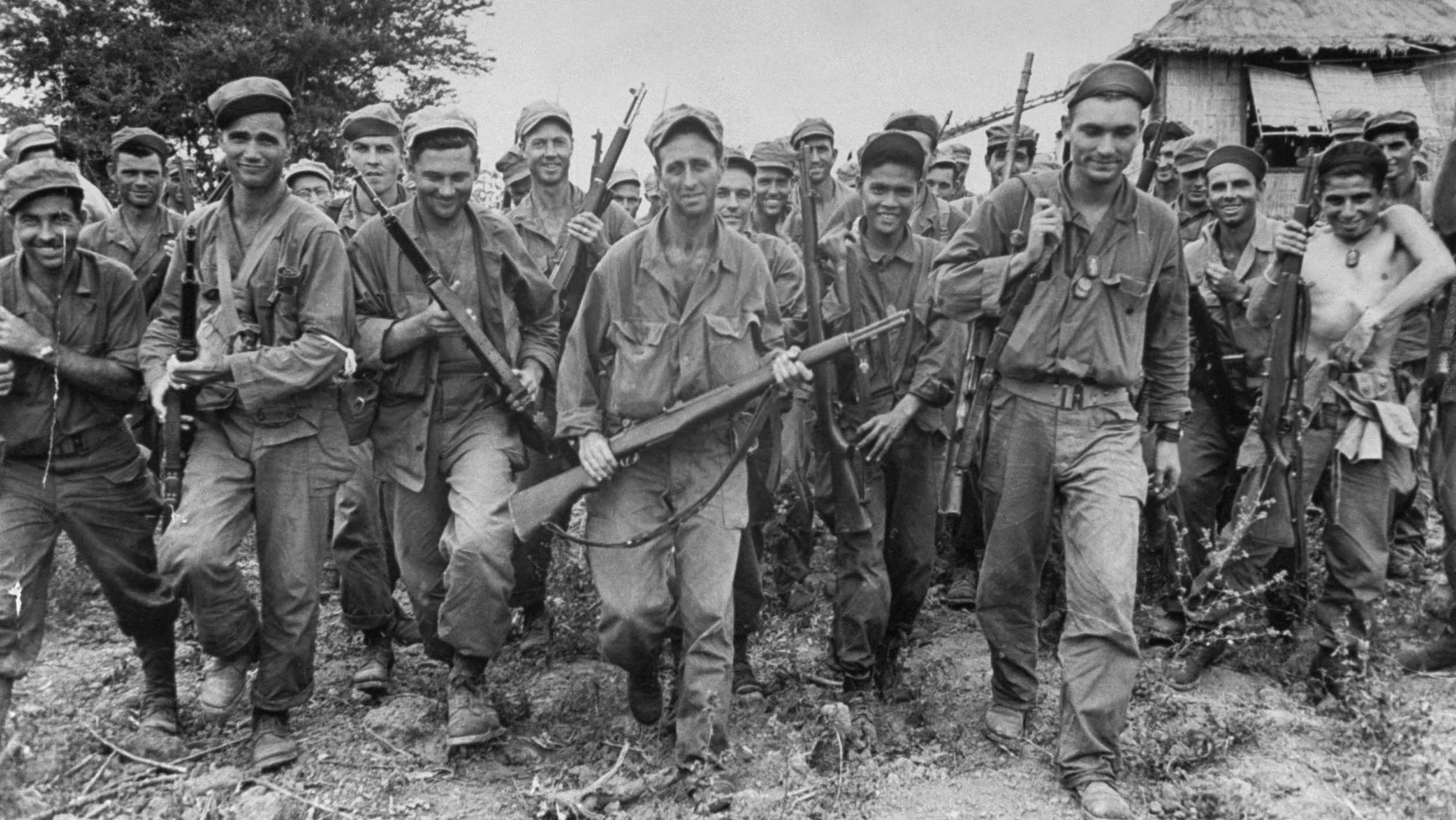 Black and white photo of a group of armed soldiers walking outdoors, some smiling, with trees and a simple wooden structure in the background.
