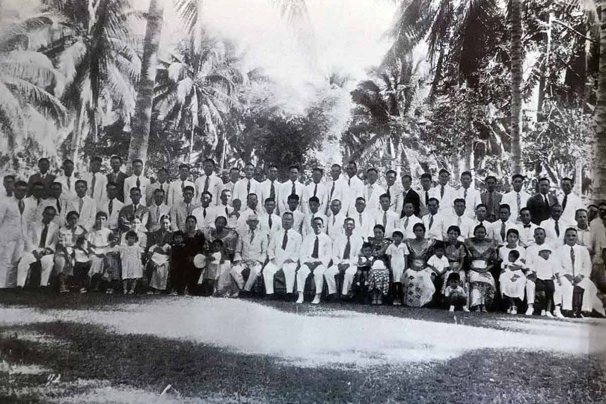 A large group of men, women, and children dressed in formal clothing, sitting and standing outdoors among palm trees in a tropical setting, in a vintage black and white photograph.