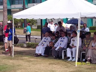 Group of people sitting under a white tent at an outdoor event, some in formal attire, with others standing nearby.