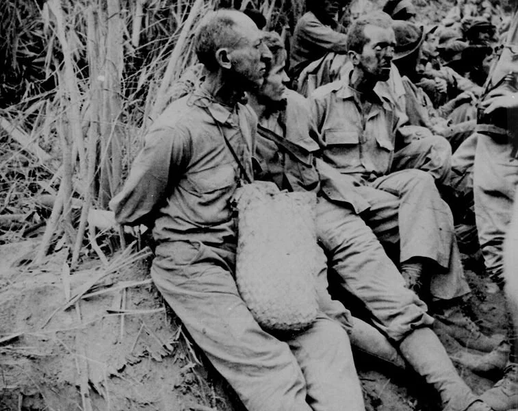 Black and white photo of soldiers sitting in a field or jungle, some with hats, appearing tired or contemplative.