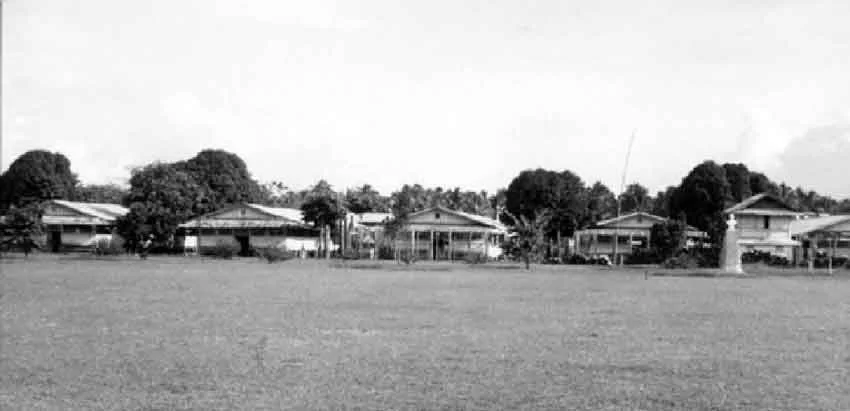 A black and white photo of several houses with trees in the background, surrounded by an open grassy area.