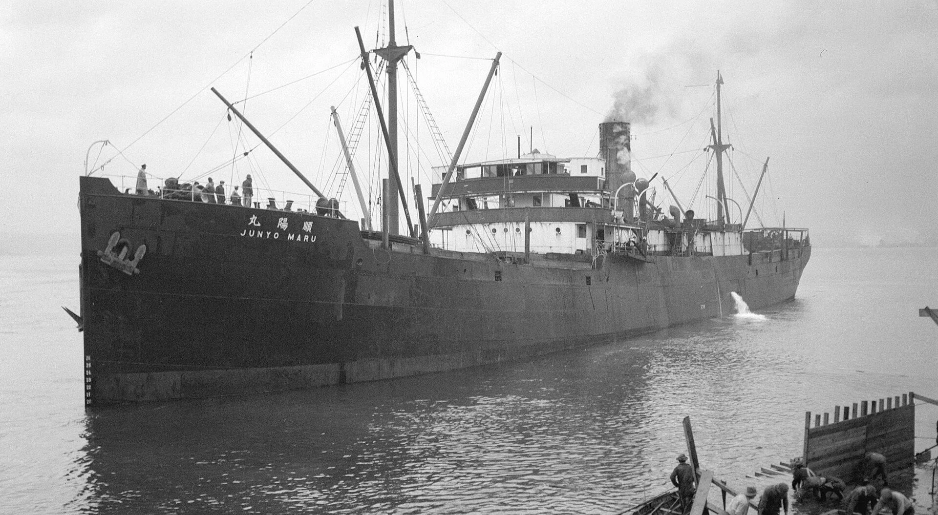 Black and white photograph of a large, old cargo ship named 'JUNYO MARU' docked at a pier, with workers on the dock and on the ship, some wearing hats, performing tasks related to loading or maintenance.