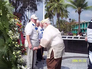 Two elderly men, one in a white suit and the other in a light-colored suit with a hat, are bowing to each other outdoors near palm trees and flowers, with to the side a woman in blue and other people in the background.