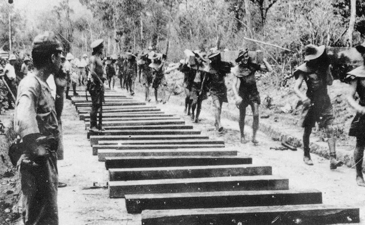 Historical black and white photo of soldiers or workers constructing a wooden bridge or pathway in a rural, wooded area.