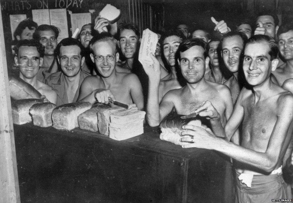 Group of shirtless men with faces of famous Americans gathered around a table of loaves of bread.