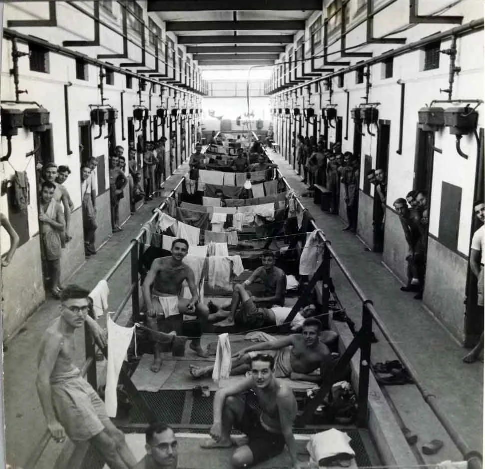 Men in a prison cell block, some sitting and lying on the beds, others standing in the doorways, with laundry hanging on a line in the center aisle.