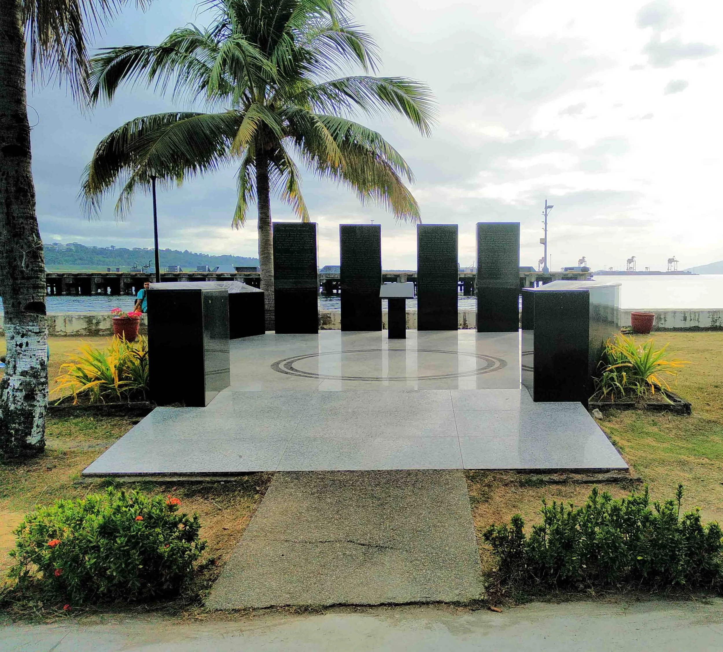 Memorial site near water with five black plaques, palm trees, potted plants, and a cloudy sky.