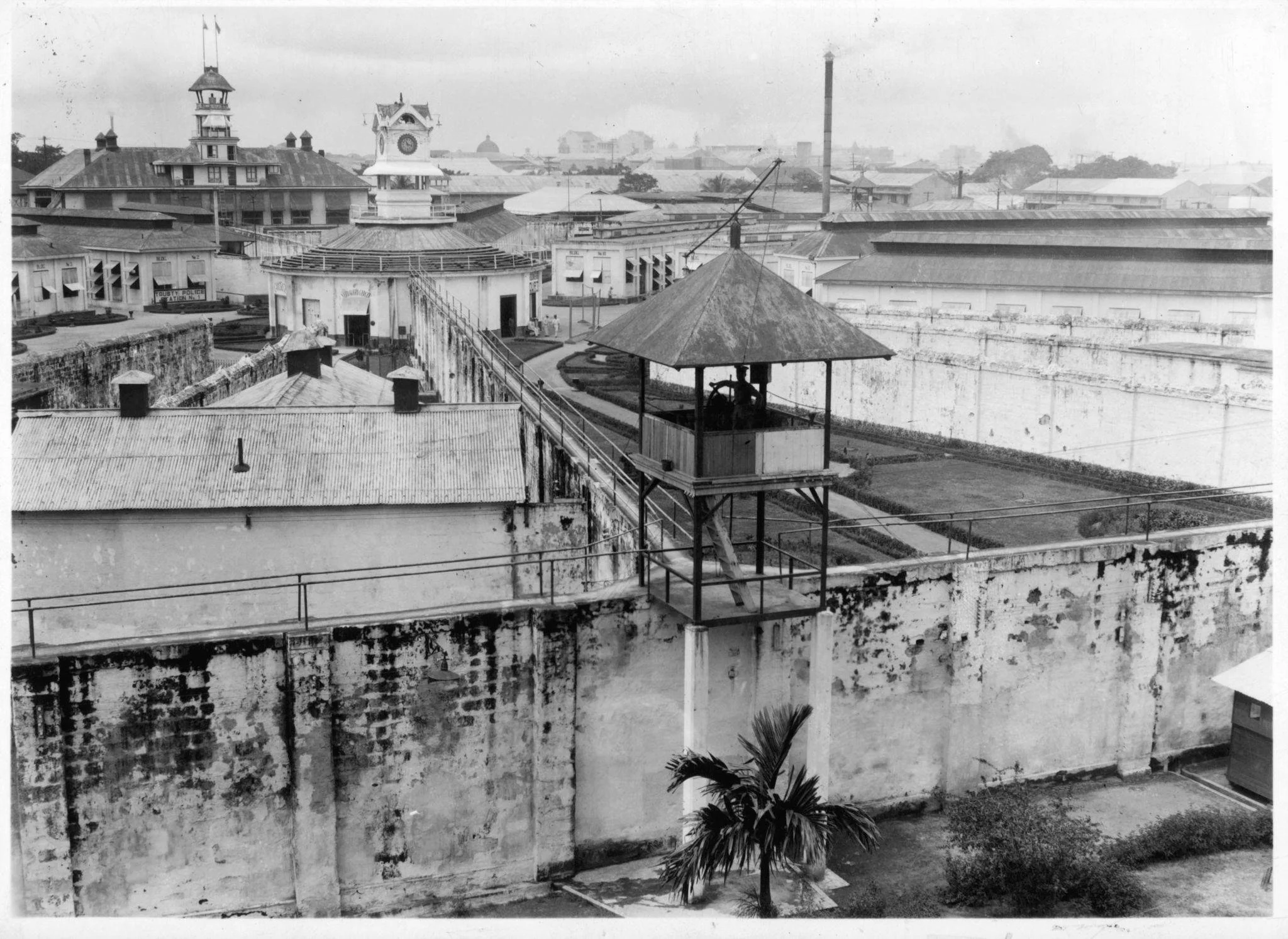 Black and white photo of a prison yard with a watchtower, surrounding walls, and multiple buildings with roofs in the background.