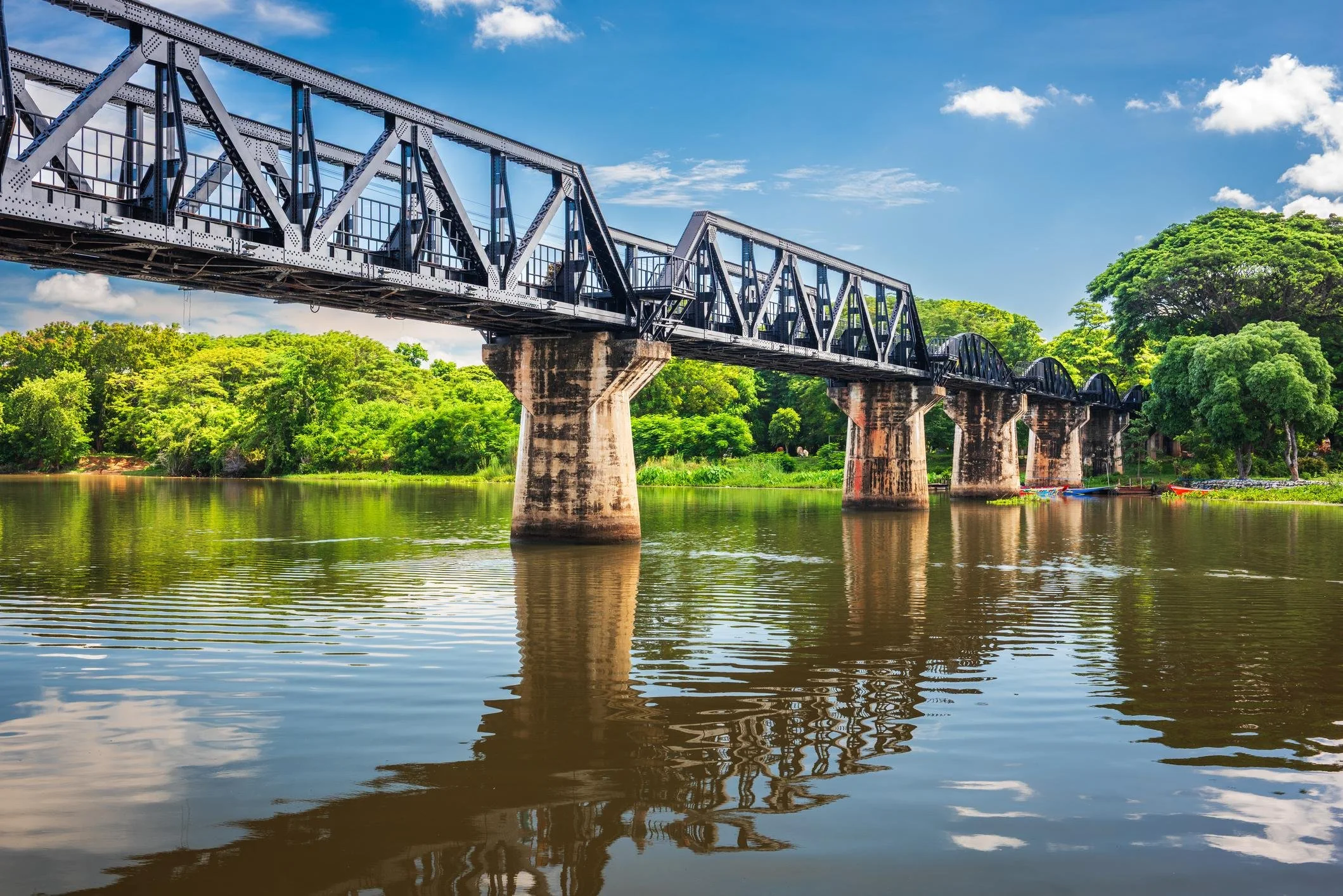 A black metal train bridge over a calm river, with lush green trees and a partly cloudy sky in the background.