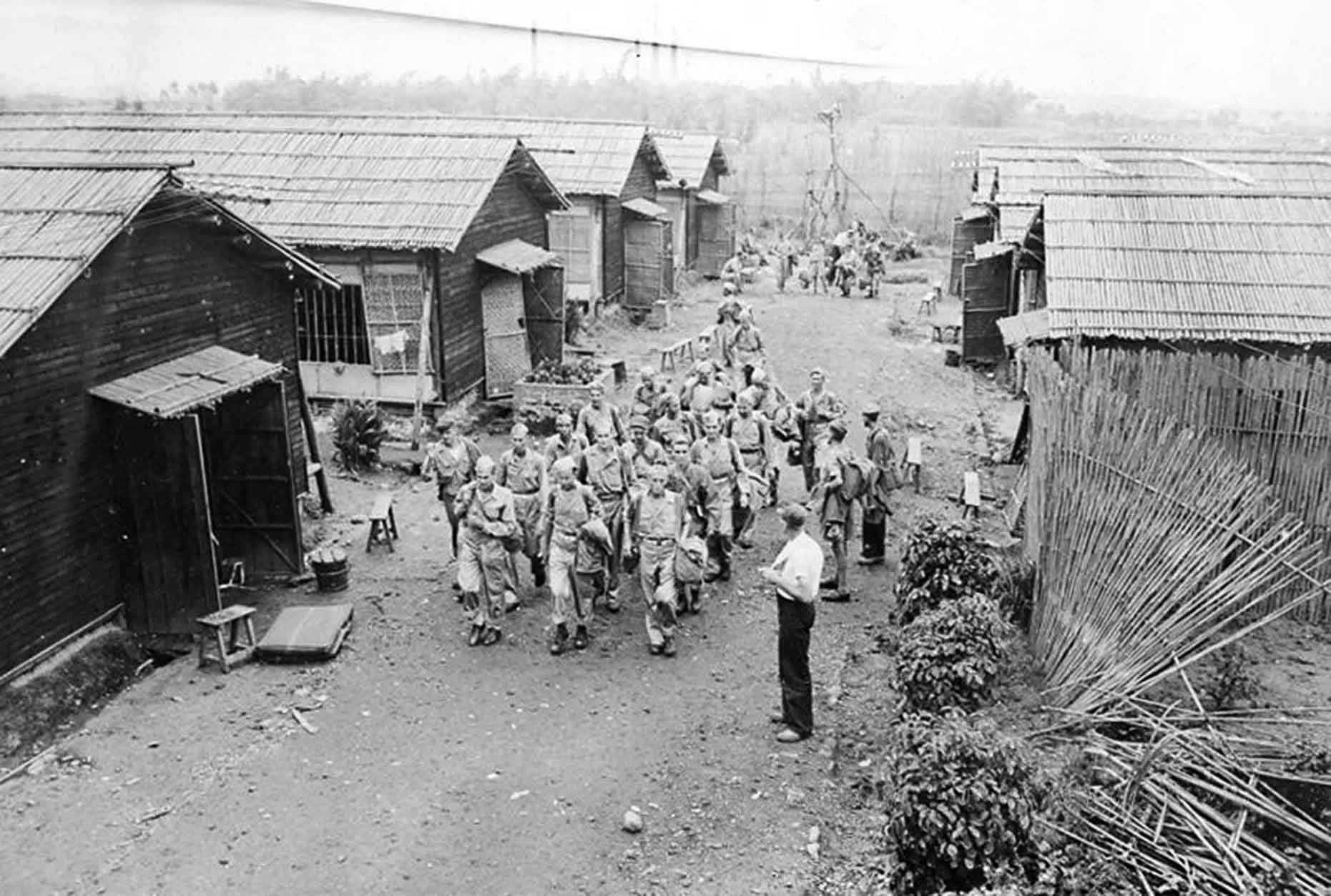 Historical black and white photograph of a group of soldiers walking through a rural village with wooden houses and fences.