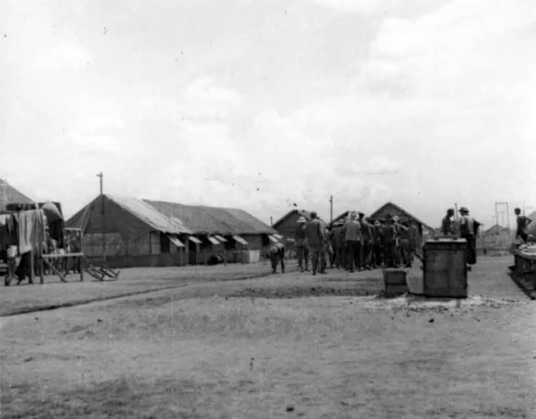 Black and white photo of a small town, with people gathered in the middle, and buildings with pitched roofs in the background under a partly cloudy sky.