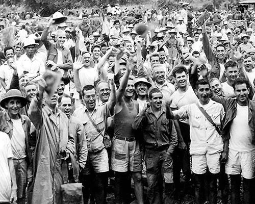 A large group of people, including men and women, gathered outdoors, many raising their hands or hats, some smiling, and a few wearing traditional clothing, suggesting a celebratory or communal event.