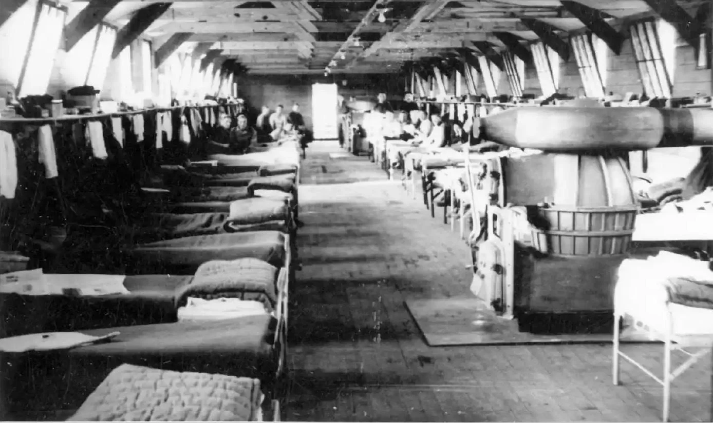Black and white photo of a dormitory or communal living space with beds along the walls and people sitting or lying on them, with a row of windows along the ceiling.